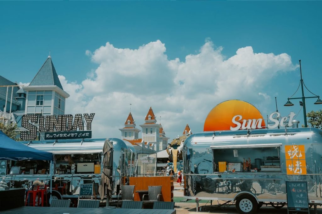 Street food carts with vibrant signage in a sunny outdoor market setting, under blue skies and fluffy clouds.